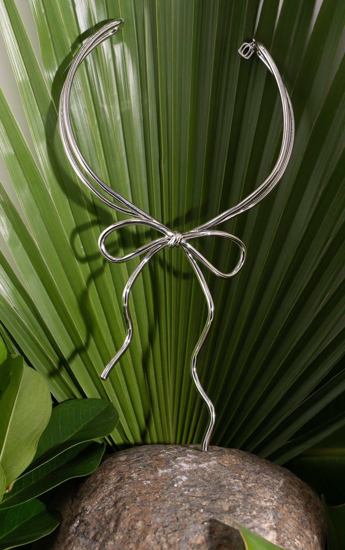A silver metal wire sculpture in the shape of a bow, displayed on a large textured stone surrounded by green tropical leaves.