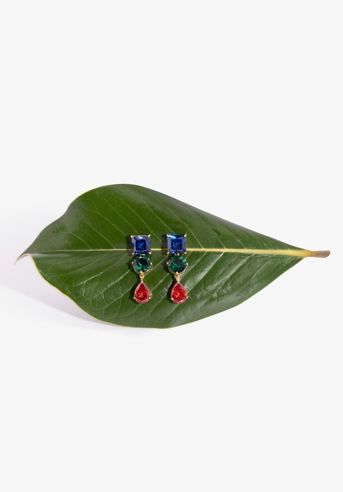A pair of gold-tone drop earrings with blue and green rectangular stones and red teardrop dangling drops, displayed on a green leaf against a white background.