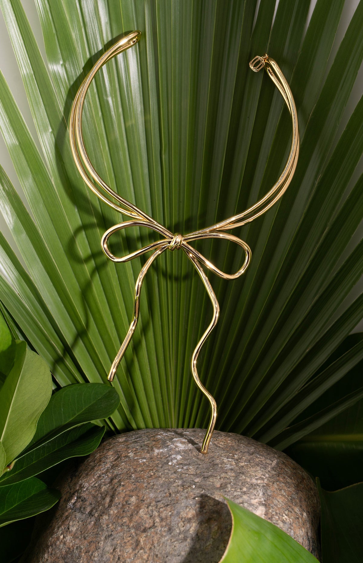 A gold wire art piece shaped like a bow with curved loops, displayed on a large brown stone surrounded by green tropical leaves.